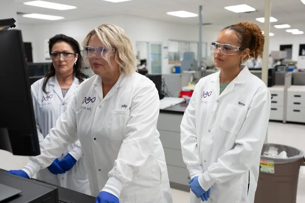Three female ACGT employees wearing protective eye glasses and white lab coats, looking at a screen