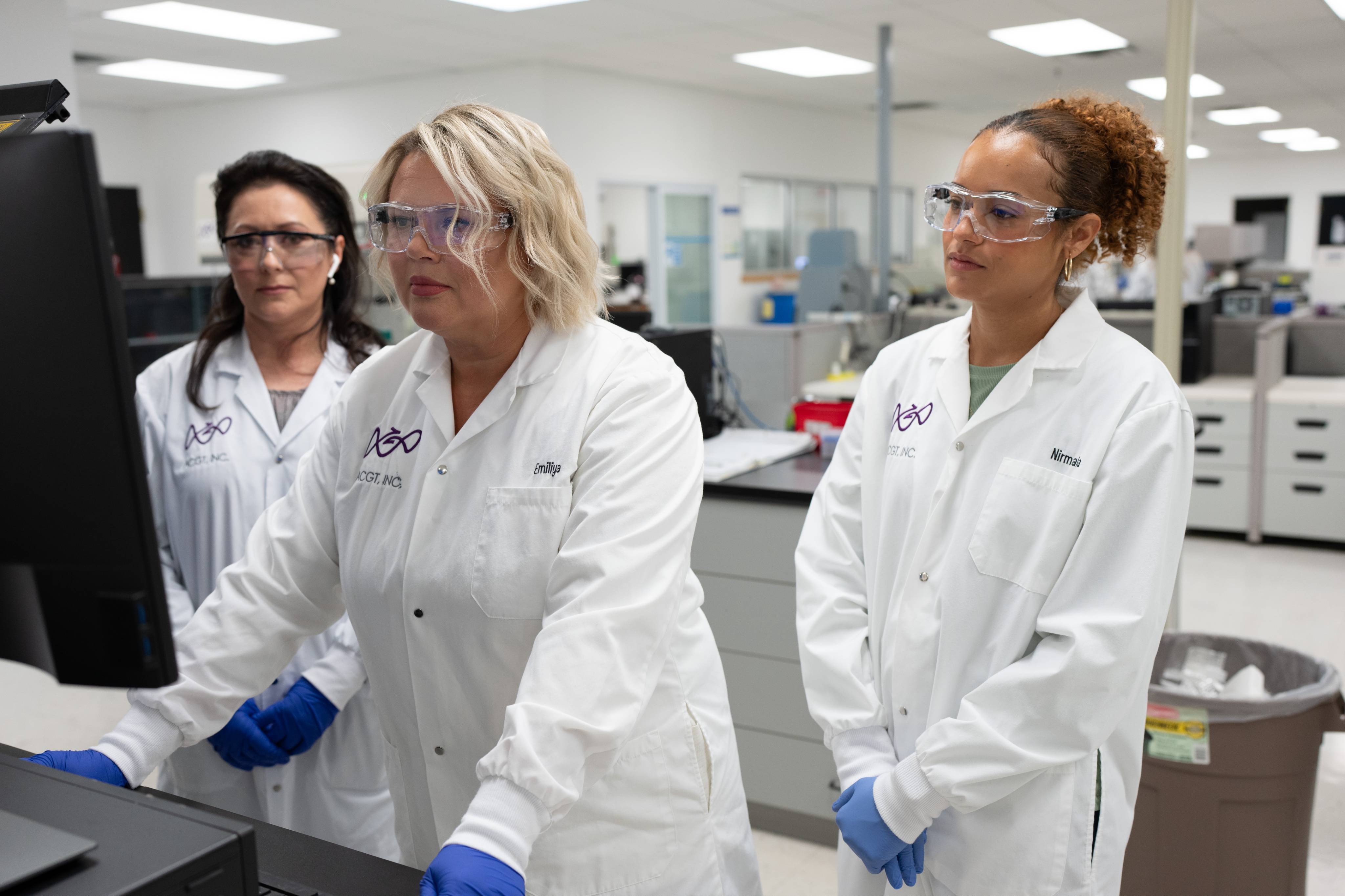 Three female ACGT employees wearing protective eye glasses and white lab coats, looking at a screen